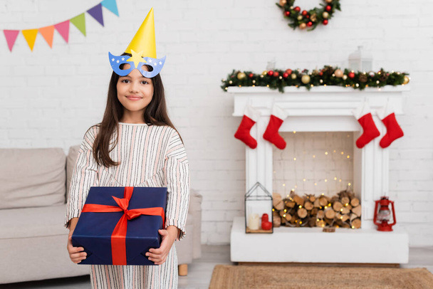Улыбающаяся девушка в маске для вечеринок и кепке держит подарок лентой во время празднования дня рождения зимой дома  - Фото, изображение