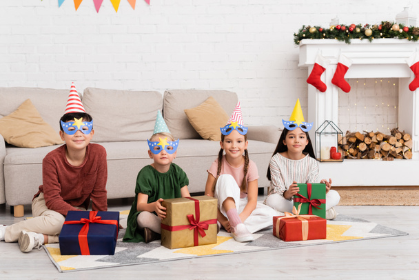 Χαμογελώντας πολυεθνικά παιδιά σε καπελάκια κόμμα και μάσκες κοιτάζοντας κάμερα κοντά δώρα κατά τη διάρκεια των γενεθλίων στο σπίτι  - Φωτογραφία, εικόνα