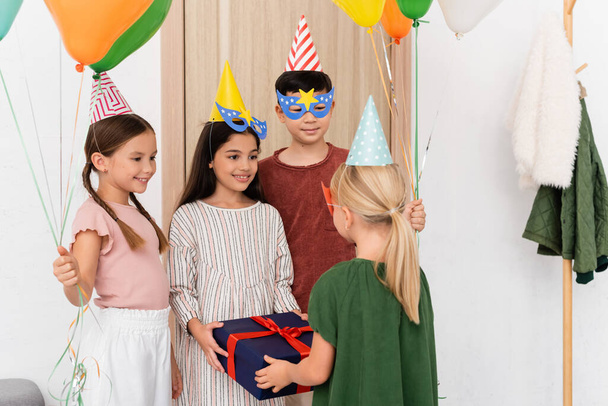 Alegre interracial niños sosteniendo regalo y globos cerca amigo en pasillo en casa  - Foto, imagen