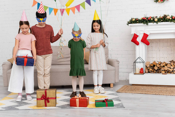Sonrientes niños multiétnicos en gorras de fiesta sosteniendo globos cerca de regalos y decoración de Navidad en casa  - Foto, imagen