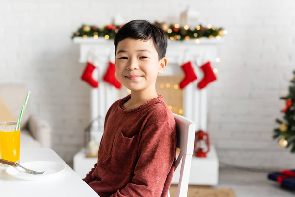 Sorridente asiatico ragazzo guardando fotocamera vicino vetro di succo d'arancia a casa  - Foto, immagini