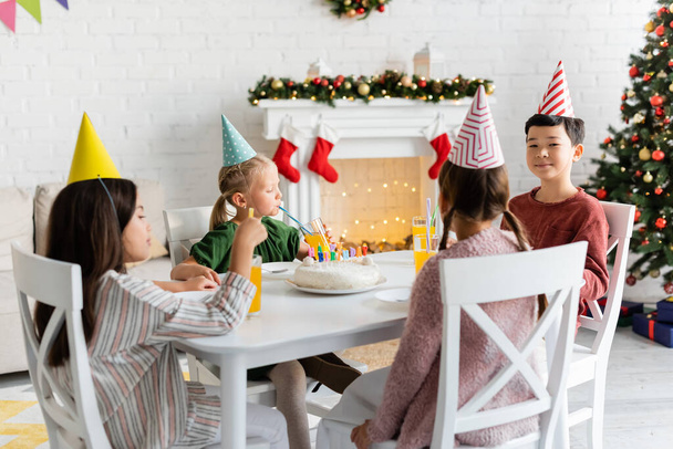 Interracial kids in party caps sitting near birthday cake and orange juice at home in winter  - Photo, image