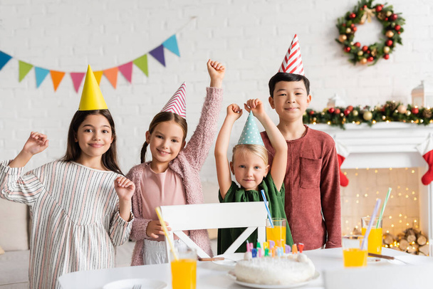 Eccitato bambini multietnici in berretti partito guardando la fotocamera vicino torta di compleanno durante la festa a casa in inverno  - Foto, immagini