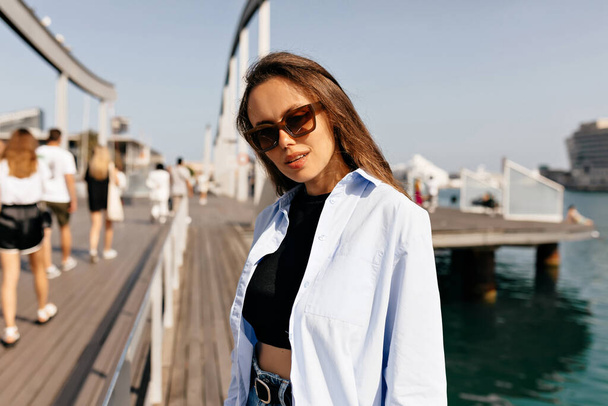 Outdoor photo of attractive caucasian young girl enjoys spending time on the pier. Brunette wears summer stylish outfit and sunglasses. Walk concept - Fotó, kép