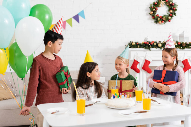 Multiethnic kids in party caps holding presents and balloons near friends and birthday cake at home  - Photo, Image