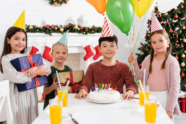 Chicas sonrientes en gorras de fiesta sosteniendo regalos y globos cerca de amigo asiático y pastel de cumpleaños en casa en invierno  - Foto, Imagen