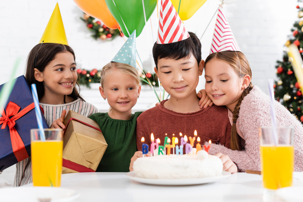 Πολυεθνικά παιδιά σε καπέλα κόμμα κρατώντας δώρα κοντά τούρτα γενεθλίων και χυμό πορτοκαλιού στο σπίτι  - Φωτογραφία, εικόνα