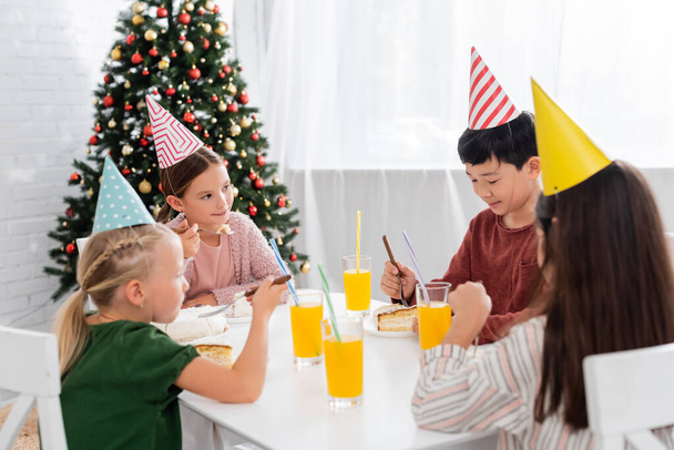Multiethnic kids in party caps celebrating birthday near cake and orange juice at home  - Photo, Image