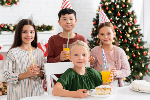 Uśmiechnięta dziewczyna patrzy na kamerę w pobliżu tortu urodzinowego i wielonarodowych przyjaciół z sokiem pomarańczowym w domu w zimie  - Zdjęcie, obraz
