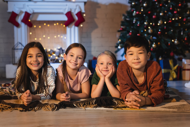 クリスマスツリーの近くの床に毛布の上に横たわっている間、カメラを見て笑顔異人種間の子供 - 写真・画像