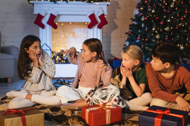 Φοβισμένο κορίτσι κάθεται κοντά σε πολυεθνικούς φίλους και παρουσιάζει κατά τη διάρκεια των Χριστουγέννων γιορτή στο σπίτι  - Φωτογραφία, εικόνα