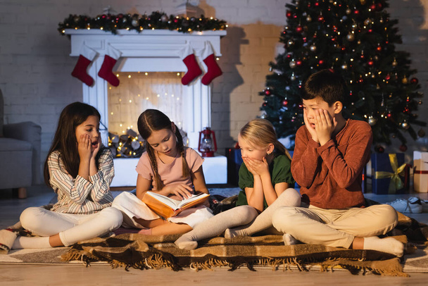 Girl reading book near scared interracial friends on blanket near christmas tree at home  - Photo, Image