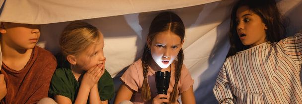Girl holding flashlight near frightened friends under blanket at home, banner  - Photo, Image
