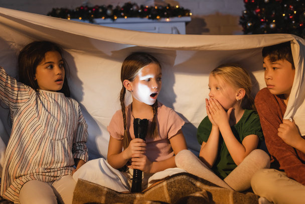 Mädchen hält Taschenlampe in der Nähe verängstigte interrassische Freunde unter Decke und Weihnachtsdekor zu Hause  - Foto, Bild