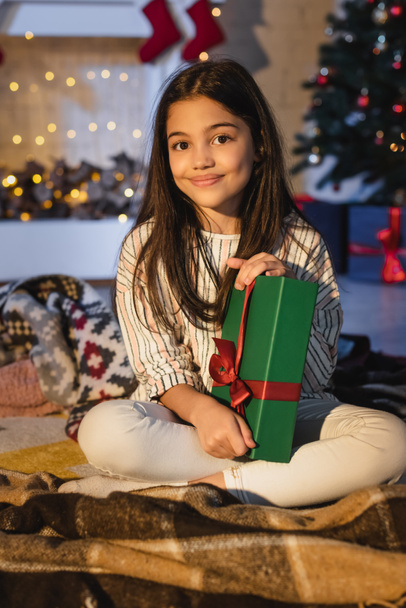 Smiling preteen girl holding gift while sitting on blanket during christmas celebration at home  - Photo, Image