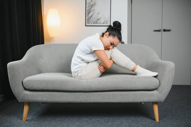 Portrait of beautiful young woman with depressed facial expression sitting on grey textile couch. Cyber bullying victim concept. Sad female in her room. Background, copy space. - 写真・画像