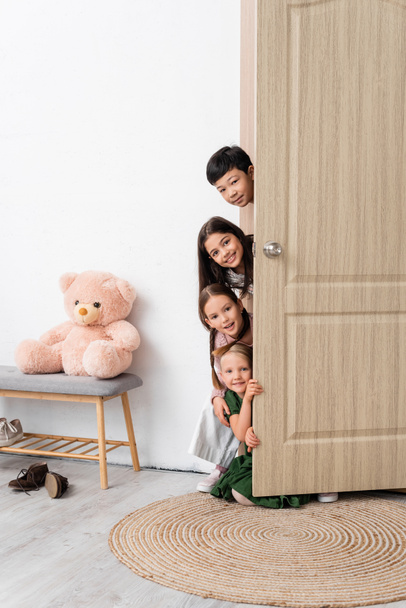 Ευτυχισμένα πολυεθνικά παιδιά που κοιτούν την κάμερα κοντά στην ανοιχτή πόρτα στο διάδρομο στο σπίτι  - Φωτογραφία, εικόνα