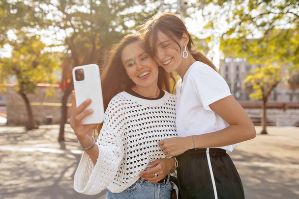 Ευτυχισμένο δύο κορίτσια με τα πόδια εξωτερική, διασκεδάζοντας μαζί και κάνοντας selfie στο smartphone i ηλιόλουστη καλοκαιρινή βραδιά στο πάρκο. Φύση, έννοια διακοπών.  - Φωτογραφία, εικόνα