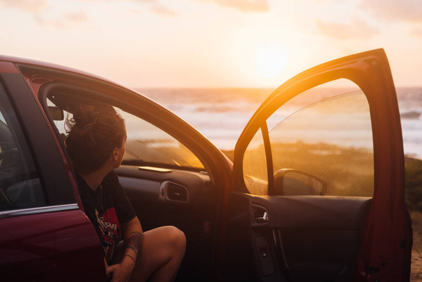 Jeune femme assise en voiture regardant le coucher du soleil à la plage - Photo, image