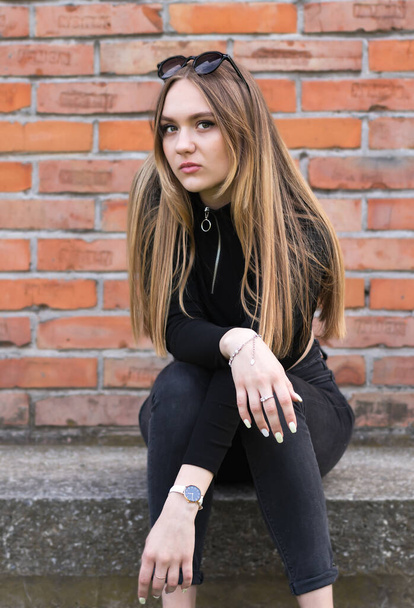 young beautiful brunette with long hair, 17 years old, student, sits against the background of a brick wall - Foto, imagen
