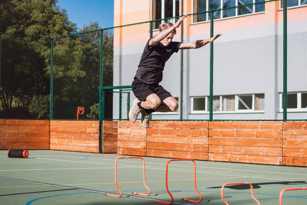 Blond boy in sportswear jumps over red obstacles to improve lower body dynamics. Plyometric training in an outdoor environment. Improve your skills. - 写真・画像