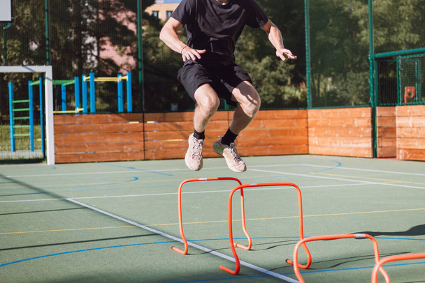 Blond boy in sportswear jumps over red obstacles to improve lower body dynamics. Plyometric training in an outdoor environment. Improve your skills. - Photo, Image