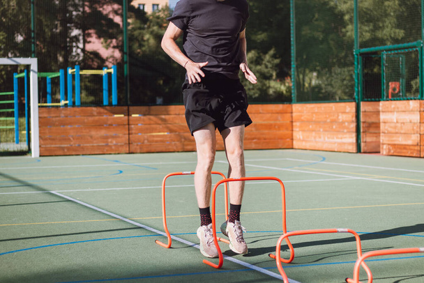 Szőke fiú sportruházatban átugrik a piros akadályokon, hogy javítsa az alsó test dinamikáját. Plyometrikus tréning szabadtéri környezetben. Javítsd a képességeidet.. - Fotó, kép