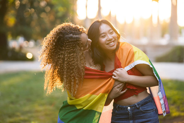 Porträt eines lesbischen Paares mit Regenbogenfahne. Vielrassiges Paar bei Sonnenuntergang. - Foto, Bild