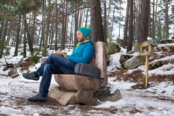 Un hombre atractivo sentado en un banco de madera mientras lee un libro sobre un bosque nevado. Concepto de encuadernación. - Foto, imagen