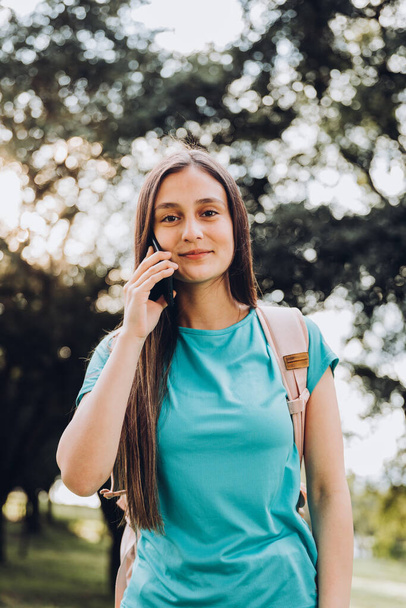 Teenage girl wearing turquoise t shirt, making a phone call with her smartphone in the park. Sun backlight. High quality photo - Foto, immagini