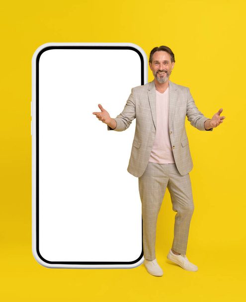 Business people, handsome middle aged businessman stand in front big smartphone with white screen spread hands. Man in white casual suit isolated on yellow background.  - Valokuva, kuva