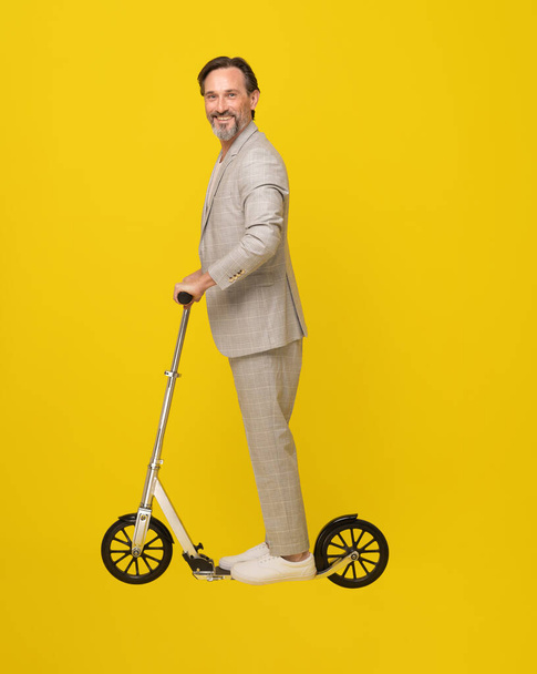 Happy middle aged man ride scooter looking sideways in white casual suit. Mobility for middle aged business man isolated on yellow background. Modern eco transport freelancer man. Business concept.  - Foto, Imagem