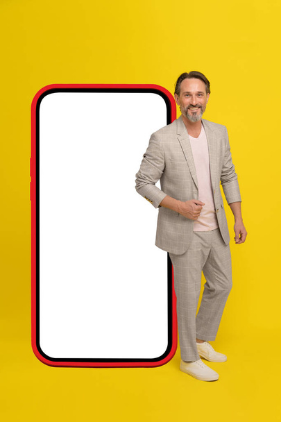 Mature business man in black suit stand in front huge smartphone in red case looking at camera isolated on yellow background. Middle aged businessman in white suit, mobile app advertisement.  - Фото, изображение