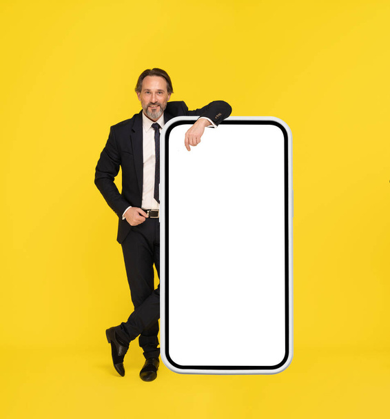 Middle aged businessman in black suit leaned on big smartphone in white case with white screen isolated on yellow background. App advertisement product placement. Man in suit.  - Foto, Bild