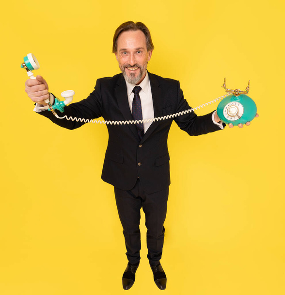 Top view. Handsome business man with old fashion telephone in hands stretched sideways looking at camera isolated on yellow background. Handsome businessman in black suit. Business concept.  - Фото, изображение
