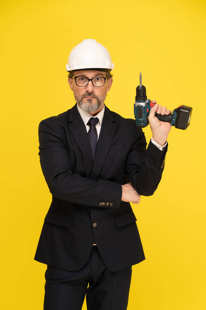 Middle aged architect businessman holding power, electric screwdriver wearing safety helmet looking at camera isolated on yellow background. Mature head of construction in black suit. - Fotó, kép