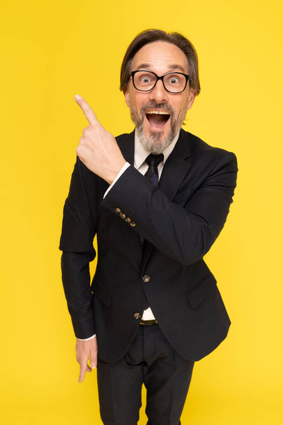 Excited happy businessman pointing finger sideways up looking at camera isolated on yellow background. Handsome mature businessman in black suit. Business concept. Mock up.  - Fotografie, Obrázek