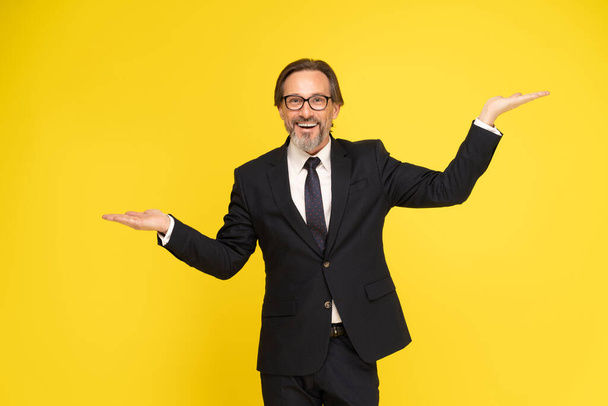 Happy middle aged business man leveling up, weighing or balancing with hands up in levels looking at camera isolated on yellow background. Handsome mature businessman in black suit. Business concept.  - Photo, image