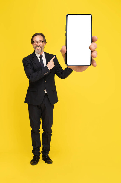 Pointing at smartphone with white screen excited middle aged businessman looking at camera isolated on yellow background. Handsome mature businessman in black suit. Business concept. Mock up.  - Foto, Bild