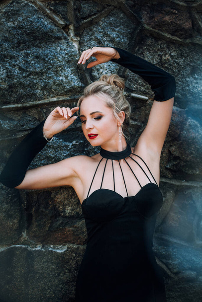 A slender beautiful woman with blond hair in a black vintage dress and expensive jewelry, stylish makeup and hairstyle, stands near the walls of an old castle. - Foto, Imagem