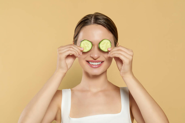 Young woman with cucumber slices on beige background. Eye skin care - Фото, изображение