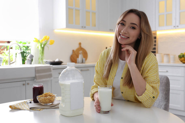 Young woman with gallon bottle of milk, glass and breakfast cereal at white table in kitchen - Photo, image