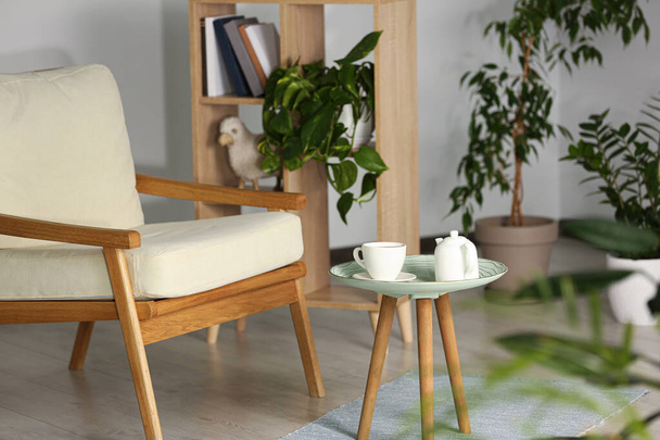 Stylish armchair, side table with cup and teapot near beautiful houseplants. Interior design - Fotoğraf, Görsel
