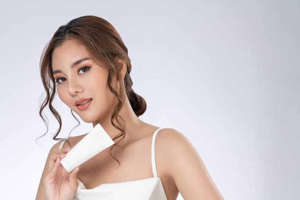 Gorgeous woman with makeup smiling holding mockup product for advertising text place, light grey background. Advertising concept for skin healthcare and beauty care products. - 写真・画像