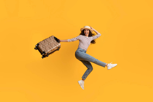 Joven viajera alegre saltando con la maleta en las manos sobre el fondo amarillo, feliz mujer milenaria emocionada en sombrero de paja llevando equipaje y divirtiéndose, disfrutando del tiempo de vacaciones, espacio de copia - Foto, imagen