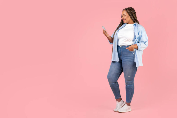 携帯電話でテキストを黒の女性とピンクのスタジオの背景に立ってインターネットを閲覧,プラスサイズの服を着てポーズ.コピースペースの近くでアプリケーションを使用して女性。モバイルの提供とコミュニケーション - 写真・画像