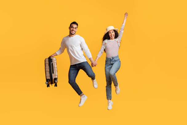 Безумное предложение. Радостная арабская пара, держась за руки и прыгая с чехлом, счастливый взволнованный ближневосточный мужчина и женщина с багажом веселясь вместе над желтым фоном, скопировать пространство - Фото, изображение