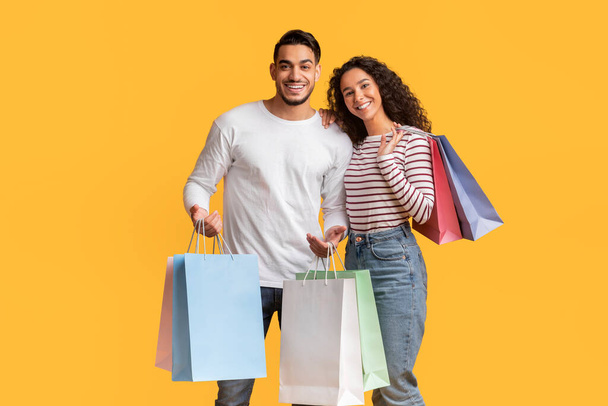 Cheerful Young Middle Eastern Couple Posing With Shopping Bags In Hands Over Yellow Background, Happy Millennial Arab Man And Woman Carrying Purchases, Enjoying Seasonal Sales, Copy Space - Φωτογραφία, εικόνα