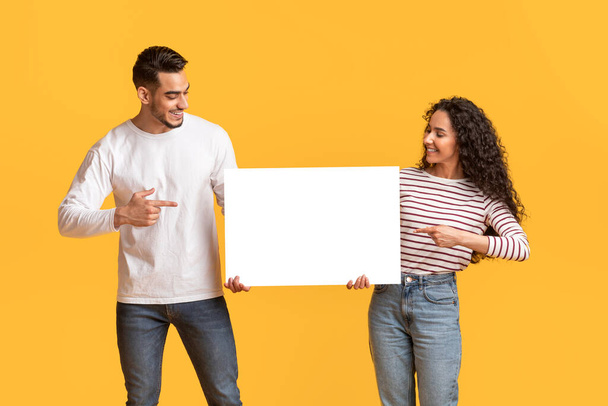 Check This. Smiling Arab Man And Woman Pointing At Blank White Placard In Their Hands, Happy Young Middle Eastern Couple Showing Free Copy Space For Design, Standing Over Yellow Background, Mockup - Zdjęcie, obraz
