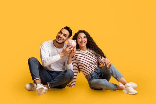 Concept d'économie. Heureux jeune couple arabe avec tirelire dans les mains posant sur fond jaune en studio, joyeux Moyen-Orient homme et femme écoutant le son des pièces, économiser de l'argent pour l'avenir - Photo, image